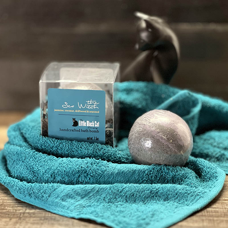 artistic arrangement of one boxed and one loose bath bomb with a towel and an art deco cat statue