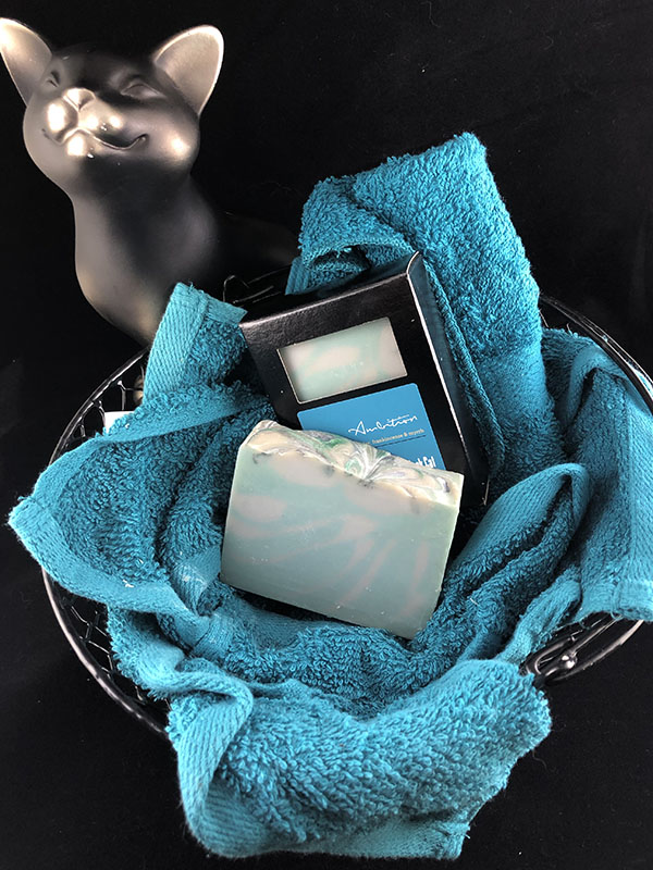 ambition soap bars, one in box one out, nested in a basket of teal towels with a sassy cat looking up at the viewer