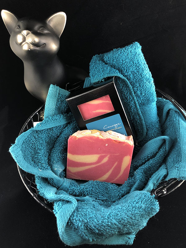 courage soap bars, one in box one out, nested in a basket of teal towels with a sassy cat looking up at the viewer