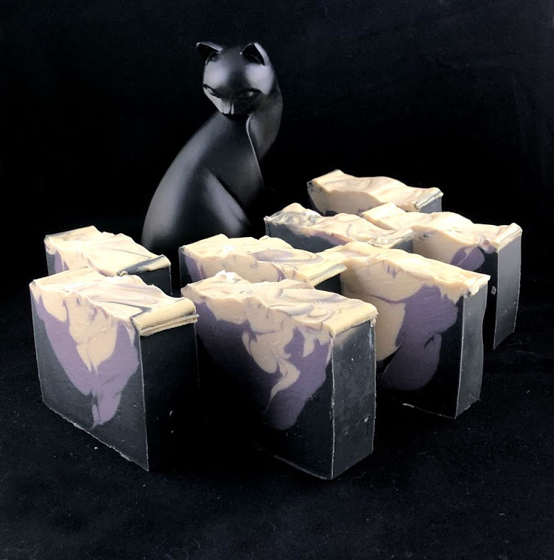 8 naked bars of black, purple-gray and white soaps arranged  standing upright with an elegant cat statue presiding in the background