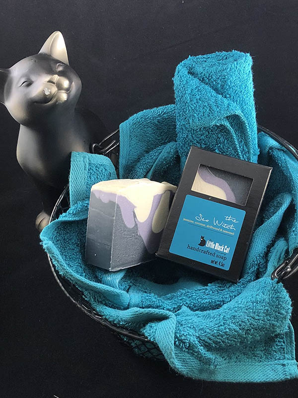 sea witch soap bars, one in box one out, nested in a basket of teal towels with a sassy cat looking up at the viewer