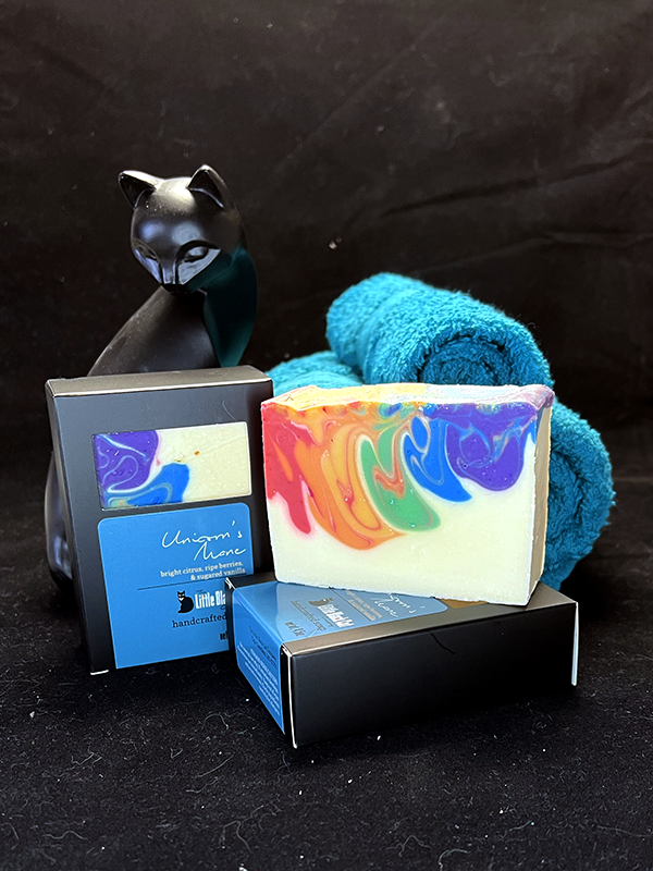 unicorn's mane soap bars, one in box one out, nested in a basket of teal towels with a sassy cat looking up at the viewer